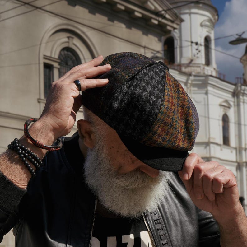 May include: A multi-colored tweed newsboy cap with a black brim. The cap features a patchwork design with houndstooth and plaid patterns in shades of brown, gray, and purple. The cap is worn by a person with a white beard.