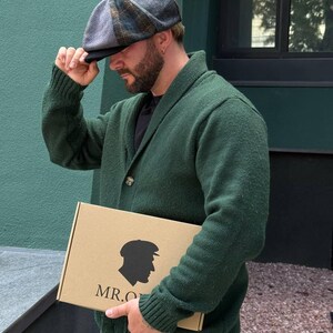 May include: A herringbone patterned newsboy cap in shades of gray, blue, and brown. The cap is being held by a person wearing a green cardigan. A cardboard box with a silhouette and the text "MR.O" is also visible.