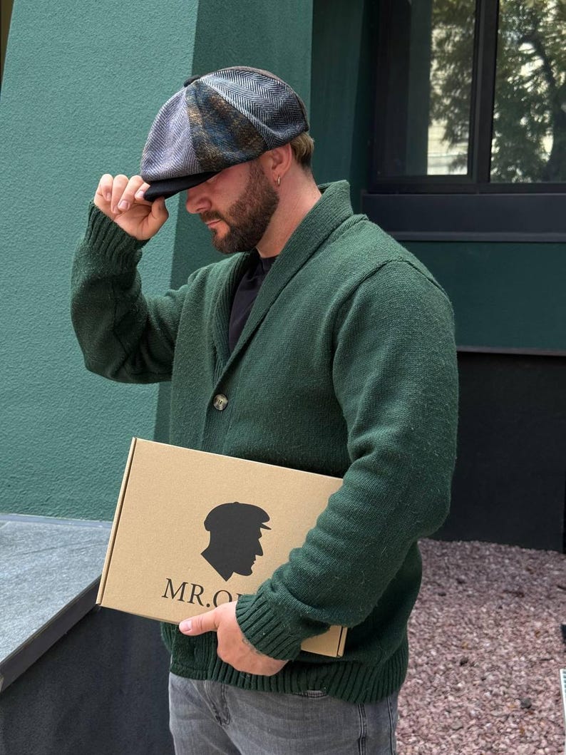 May include: A herringbone patterned newsboy cap in shades of gray, blue, and brown. The cap is being held by a person wearing a green cardigan. A cardboard box with a silhouette and the text "MR.O" is also visible.