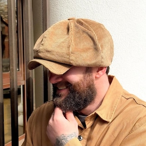 Może przedstawiać: Beżowa czapka typu newsboy wykonana z sztruksu, posiadająca wielopanelową koronę, guzik na środku góry oraz krótki, sztywny daszek. Materiał charakteryzuje się wyraźną pionową prążkowaną fakturą pokrywającą całą powierzchnię nakrycia głowy.