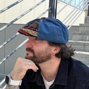 Può includere: Un berretto newsboy a otto spicchi che combina tessuto denim blu e un pannello in tessuto con motivi geometrici neri, arancioni e crema. Il berretto presenta una corona arrotondata, una visiera corta e rigida e un piccolo bottone in cima.