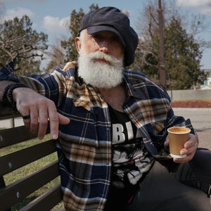 May include: A man wearing a blue and yellow plaid shirt, a black newsboy cap, and a black t-shirt with the word "BIG" printed on it. He is sitting on a wooden bench and holding a brown paper cup of coffee.