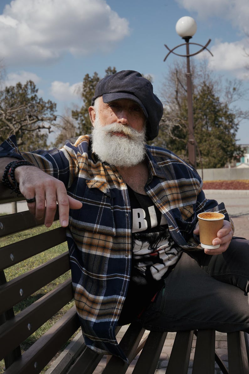 May include: A man wearing a blue and yellow plaid shirt, a black newsboy cap, and a black t-shirt with the word "BIG" printed on it. He is sitting on a wooden bench and holding a brown paper cup of coffee.