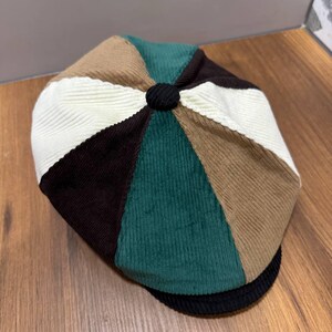May include: A multi-colored corduroy flat cap featuring a patchwork design of eight triangular panels in cream, tan, dark brown, and forest green. The cap has a small black fabric-covered button at the center crown and a black corduroy brim.