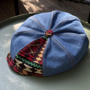 May include: A denim newsboy cap with a patterned brim. The cap is primarily blue denim with a patterned section featuring red, green, and yellow geometric designs. The cap has a button on top and is stitched with tan thread.