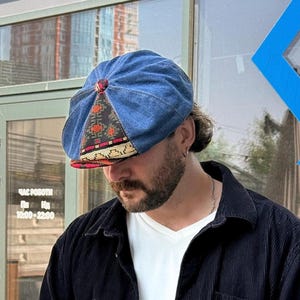 May include: A blue denim flat cap featuring a multi-paneled design with a central triangular insert of black fabric decorated with a repeating red and gold geometric pattern, finished with a matching fabric-covered button at the crown.