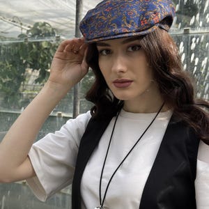 May include: A blue flat cap featuring an intricate orange and gold floral pattern, paired with a white t-shirt, a black waistcoat, and a long black cord necklace with a large, smooth, silver-toned heart-shaped pendant hanging at the centre.