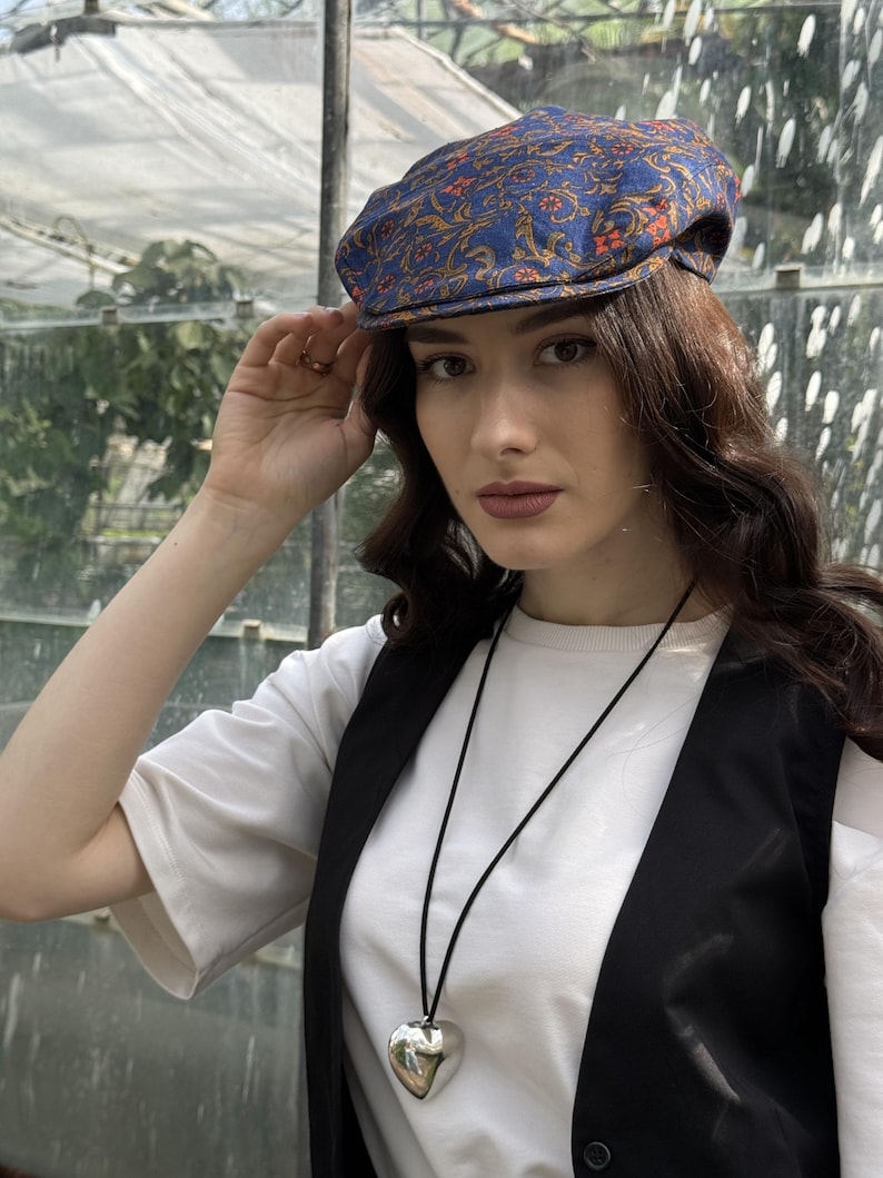 May include: A blue flat cap featuring an intricate orange and gold floral pattern, paired with a white t-shirt, a black waistcoat, and a long black cord necklace with a large, smooth, silver-toned heart-shaped pendant hanging at the centre.