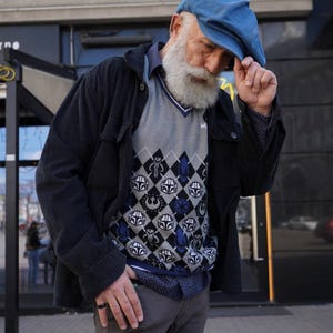 May include: A man wearing a blue denim cap, a black jacket, a gray sweater with a blue and white pattern, and gray pants. The sweater has a pattern of various Star Wars characters.