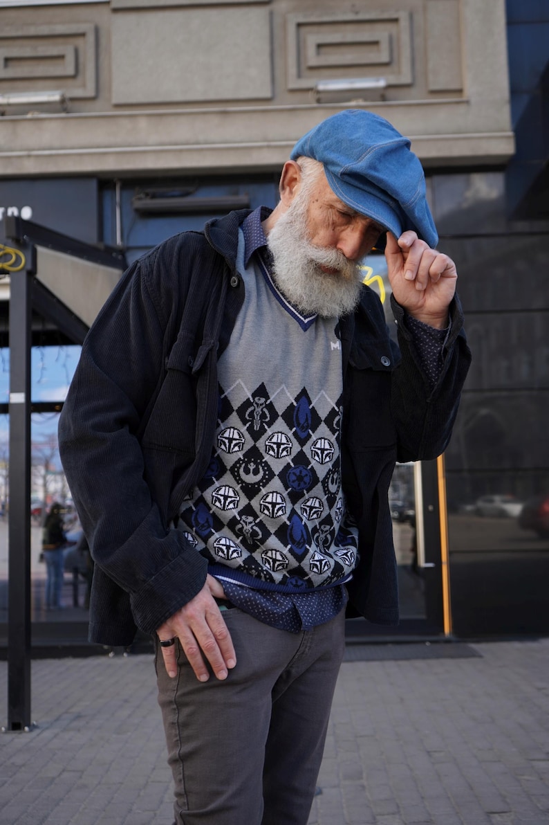 May include: A man wearing a blue denim cap, a black jacket, a gray sweater with a blue and white pattern, and gray pants. The sweater has a pattern of various Star Wars characters.