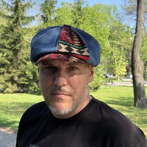 May include: A blue denim newsboy cap with a patterned brim featuring red, green, and black geometric designs. The cap has a rounded crown and a short, stiff brim. The hat is a stylish accessory.
