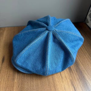May include: A blue denim newsboy cap with a rounded crown and a short, stiff brim. The cap features visible white stitching and a button detail at the top. The hat is made of a sturdy denim material.