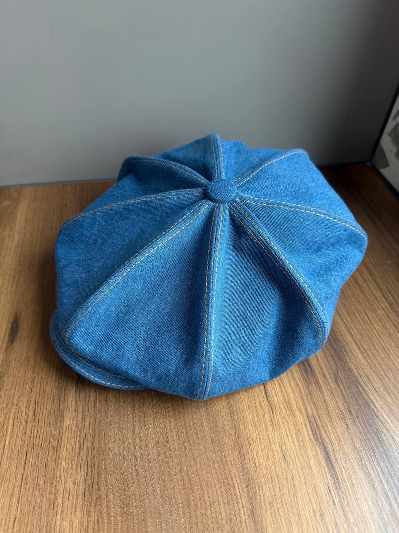 May include: A blue denim newsboy cap with a rounded crown and a short, stiff brim. The cap features visible white stitching and a button detail at the top. The hat is made of a sturdy denim material.