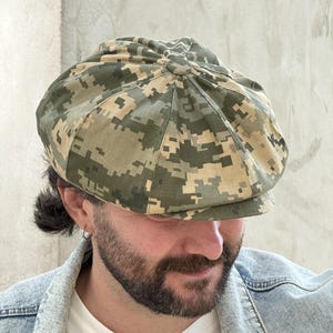 May include: A newsboy cap featuring a digital camouflage pattern in shades of olive green, dark green, tan, and brown. The cap is constructed from multiple fabric panels joined at a central button on the crown, with a short, stiffened brim.