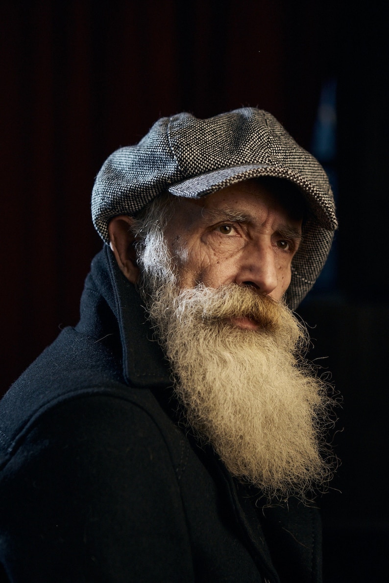 May include: A close-up portrait of an older man with a long white beard. He is wearing a black jacket and a grey newsboy cap. The man is looking off to the side.