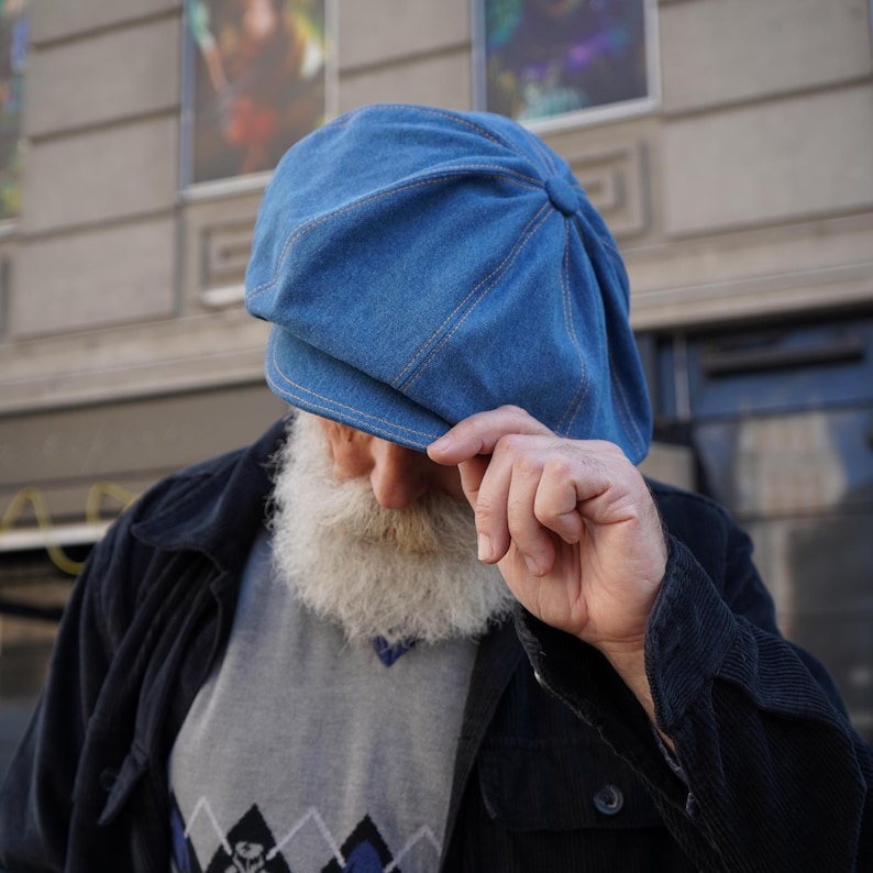 May include: A blue denim newsboy cap with a rounded crown and a small brim. The cap has visible stitching and a button detail at the top. The cap is worn by a person wearing a gray sweater and a black jacket.
