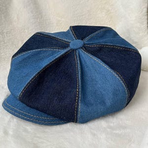 May include: A denim newsboy cap in shades of blue. The cap features a patchwork design with contrasting panels and visible stitching. The brim is a solid blue, and the top has a button detail. The hat is set against a white, textured background.