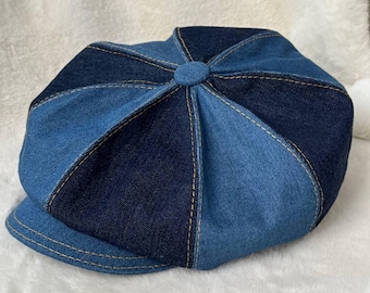 May include: A denim newsboy cap in shades of blue. The cap features a patchwork design with contrasting panels and visible stitching. The brim is a solid blue, and the top has a button detail. The hat is set against a white, textured background.