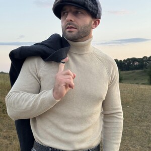 May include: A beige turtleneck sweater and patterned pants are worn with a dark gray tweed newsboy cap. A black coat is draped over the shoulder. The outfit is completed with a black belt.