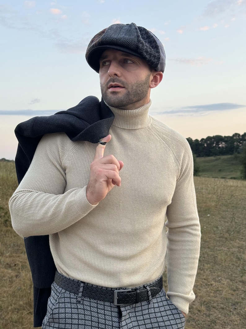 May include: A beige turtleneck sweater and patterned pants are worn with a dark gray tweed newsboy cap. A black coat is draped over the shoulder. The outfit is completed with a black belt.