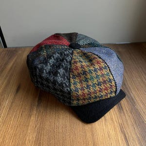 May include: A patchwork newsboy cap with a black brim. The cap features a variety of patterned fabrics in shades of red, blue, green, and gray. The cap is made of a soft, textured material.