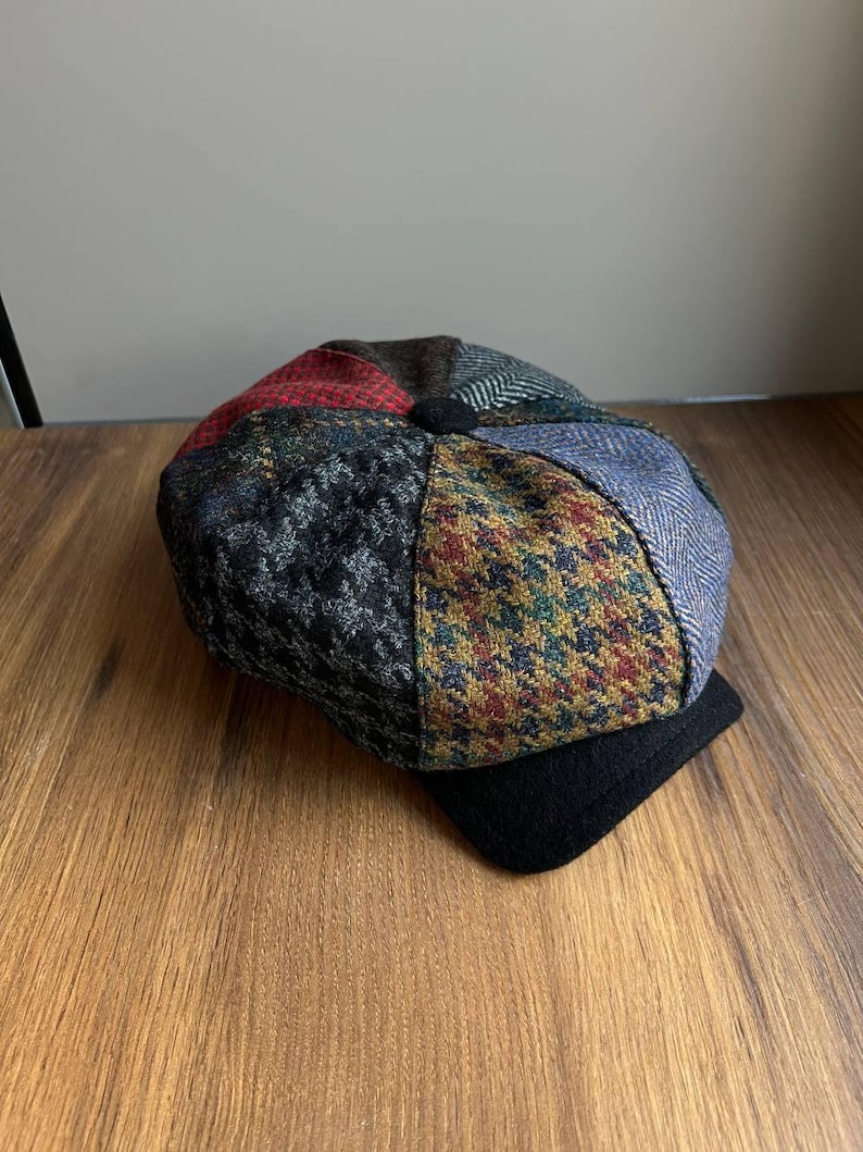 May include: A patchwork newsboy cap with a black brim. The cap features a variety of patterned fabrics in shades of red, blue, green, and gray. The cap is made of a soft, textured material.