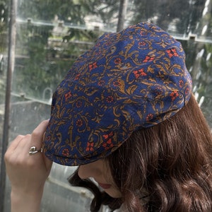 May include: A flat cap featuring a deep blue base colour with an intricate, repeating pattern of swirling golden-brown vines and small, vibrant orange floral accents. The cap has a structured, rounded crown and a short, stiff brim at the front.