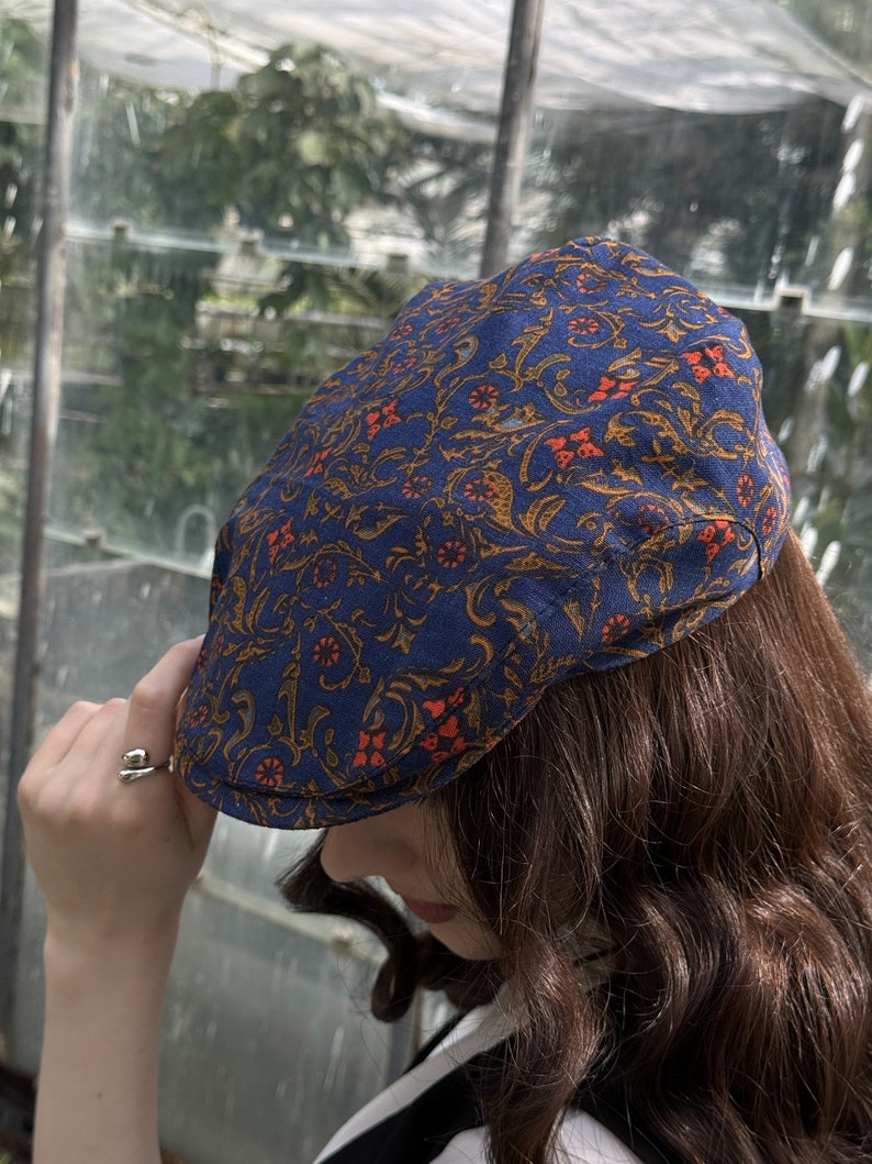 May include: A flat cap featuring a deep blue base colour with an intricate, repeating pattern of swirling golden-brown vines and small, vibrant orange floral accents. The cap has a structured, rounded crown and a short, stiff brim at the front.