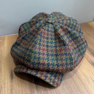 Peut inclure: Casquette de style gavroche en tissu à motif pied-de-poule multicolore. Le chapeau présente un mélange de vert, rouge, bleu et marron. Il a une couronne arrondie et une petite visière rigide. Le tissu est probablement en laine ou en mélange de laine.