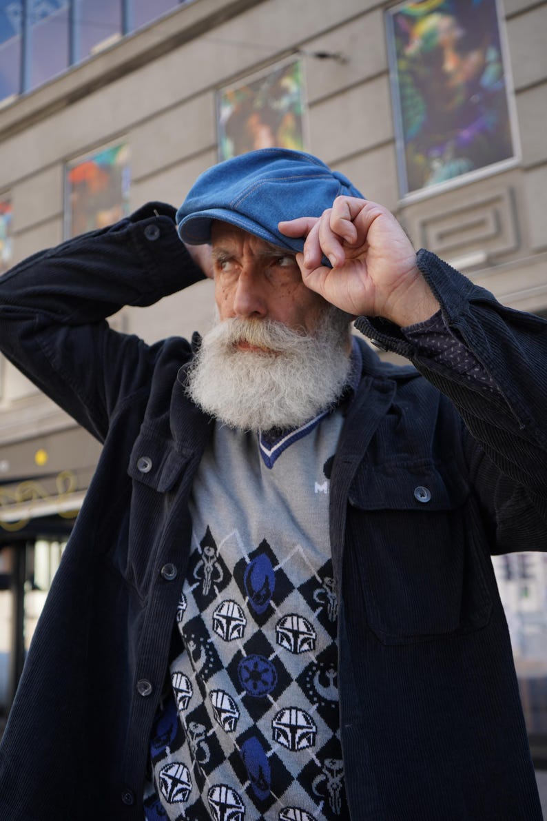 May include: A man wearing a blue denim cap, a black corduroy jacket, and a gray sweater with a black and blue geometric pattern featuring Star Wars characters.