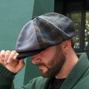 May include: A multi-toned newsboy cap with a black brim. The cap features a patchwork design with gray, blue, and brown herringbone and plaid patterns. The cap is made of a textured fabric.