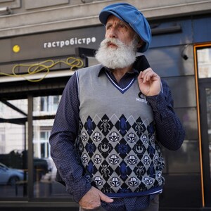 May include: A man wearing a blue newsboy cap and a gray vest with a blue and white pattern featuring Star Wars characters. The vest has a white collar and a blue trim. The man has a long white beard and is wearing a blue button-down shirt with white polka dots.