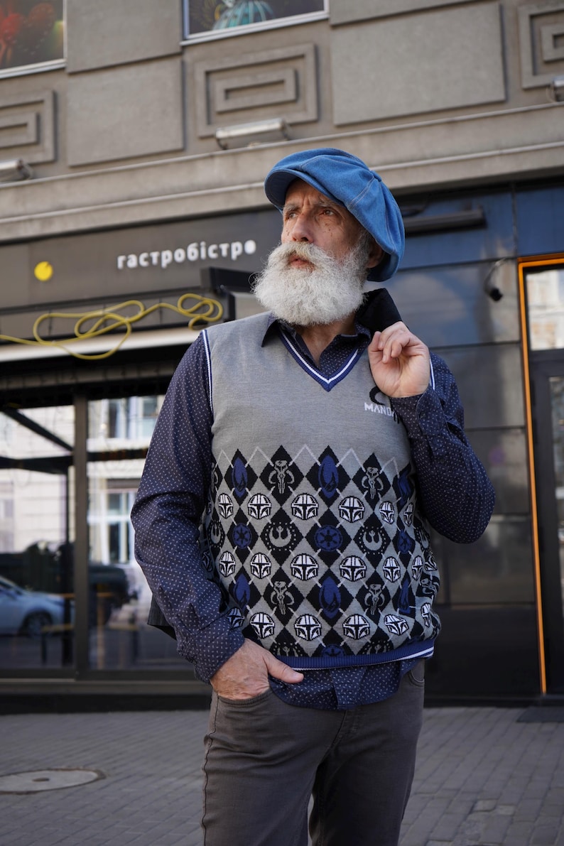 May include: A man wearing a blue newsboy cap and a gray vest with a blue and white pattern featuring Star Wars characters. The vest has a white collar and a blue trim. The man has a long white beard and is wearing a blue button-down shirt with white polka dots.