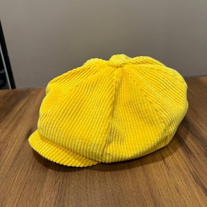 May include: A bright yellow corduroy newsboy cap. The cap has a rounded crown and a short, stiff brim. The fabric has a ribbed texture. The hat is sitting on a wooden surface.