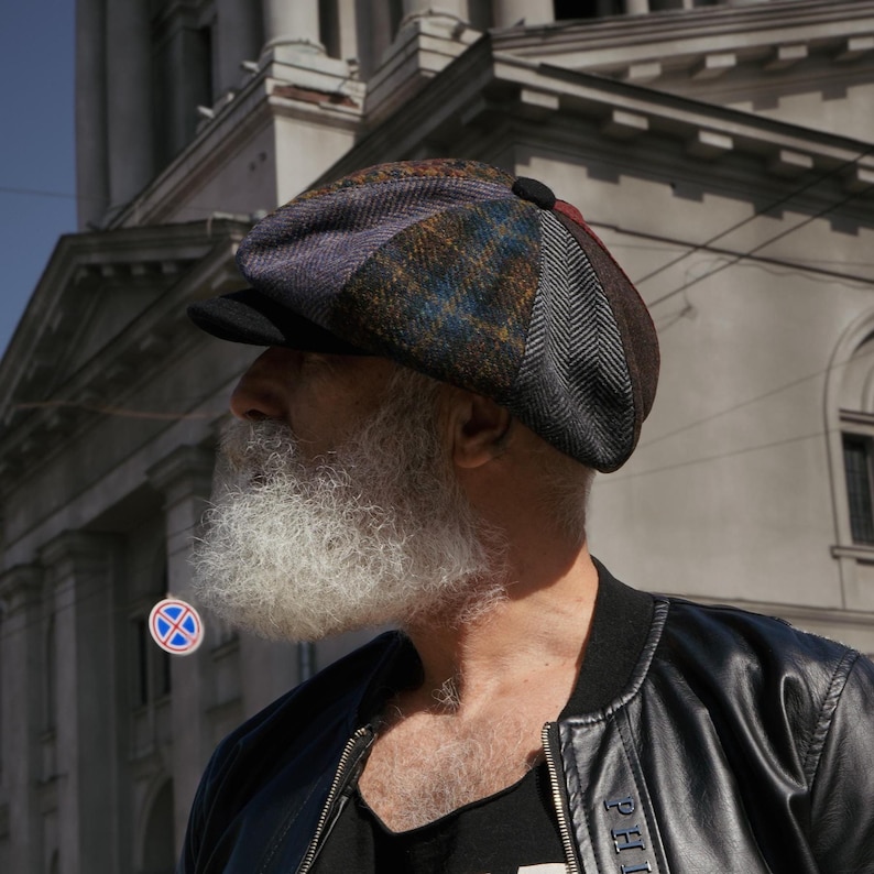 May include: A multi-colored newsboy cap with a black brim. The cap features a patchwork design with various patterns and colors, including blue, brown, and purple. The cap is worn by a person with a long, white beard and a black jacket.