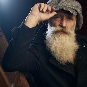 May include: A portrait of a person wearing a gray tweed newsboy cap and a black coat. The person has a long, white beard and mustache. The person is touching the cap with their hand. The background is dark.