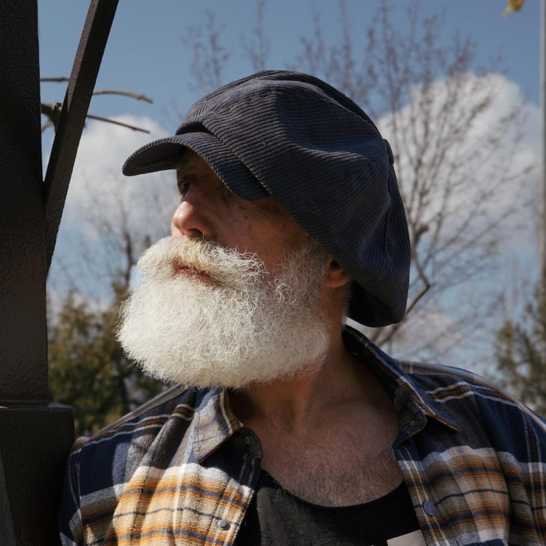 May include: A man with a long white beard wearing a blue corduroy newsboy cap and a plaid shirt.