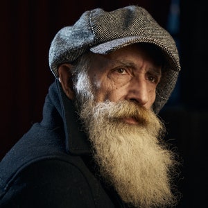 May include: A portrait of an older person with a long, white beard and a gray and black patterned cap. The person is wearing a dark coat. The background is dark, emphasizing the subject's features.