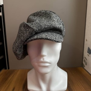 May include: A black and white tweed newsboy cap. The cap is displayed on a white mannequin head. The hat has a rounded crown and a small, stiff brim. The fabric has a textured appearance.