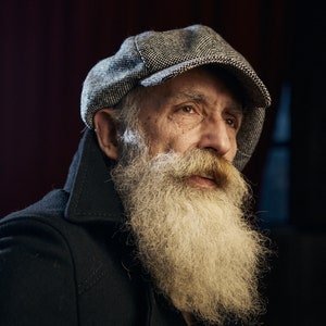 May include: A portrait of a person with a long, white beard and a tweed newsboy cap. The person is wearing a dark coat. The background is a dark red.