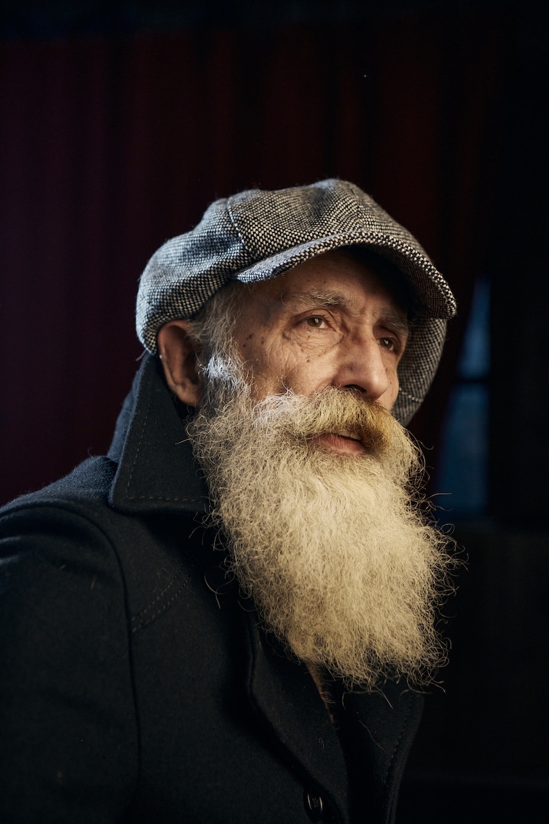 May include: A portrait of a person with a long, white beard and a tweed newsboy cap. The person is wearing a dark coat. The background is a dark red.