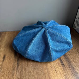 May include: A blue denim beret with white stitching. The beret has a round shape with a central button detail and a series of stitched panels radiating outwards. The hat is made of a medium-wash denim fabric.
