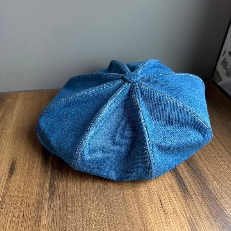 May include: A blue denim beret with white stitching. The beret has a round shape with a central button detail and a series of stitched panels radiating outwards. The hat is made of a medium-wash denim fabric.