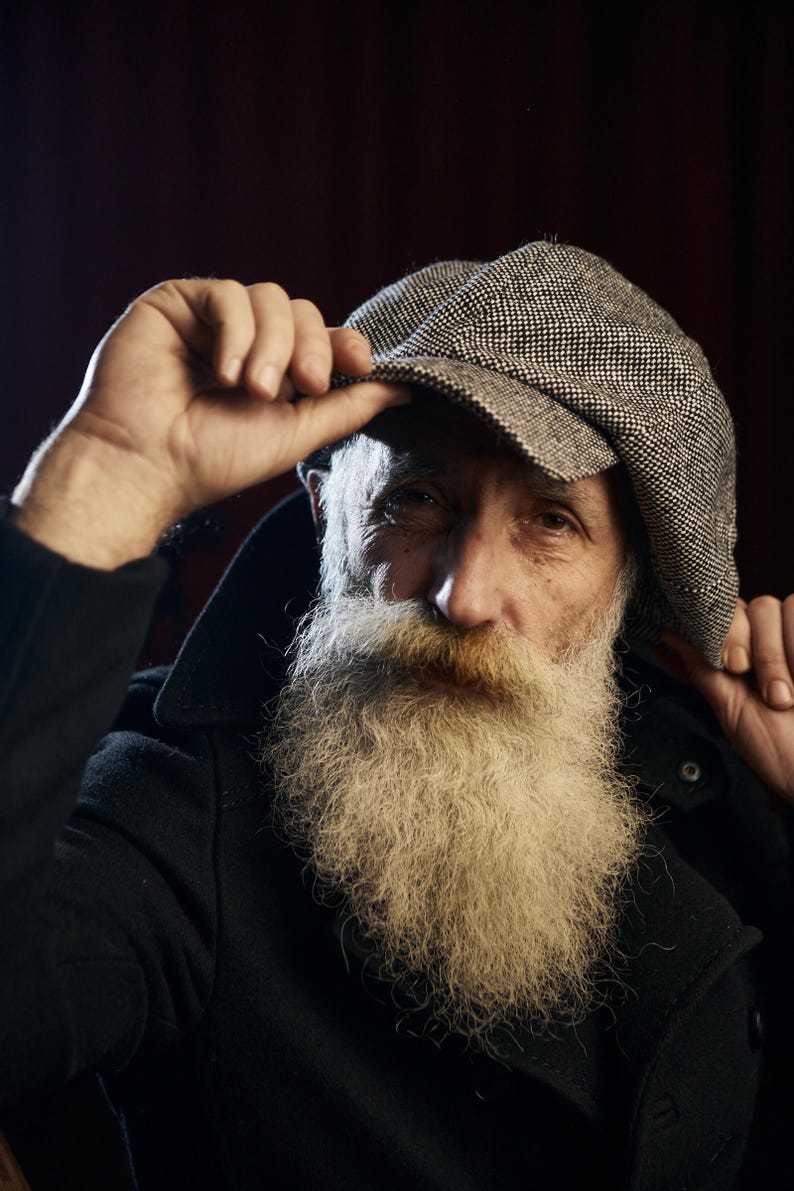 May include: A portrait of a person wearing a tweed newsboy cap. The cap is gray and white. The person has a long, white beard and is wearing a black coat. The background is a dark red.