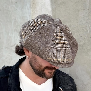 May include: A brown newsboy cap featuring a multi-panel construction with a herringbone weave pattern and a subtle plaid overlay in white and yellow. The cap has a rounded, structured crown and a short, stiff brim extending from the front.