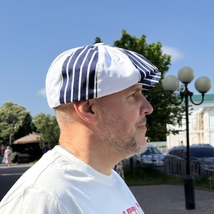 May include: A newsboy cap featuring a pattern of alternating white and navy blue vertical stripes. The cap has a rounded, multi-paneled crown construction with a short, stiff brim at the front, designed to sit securely on the head.
