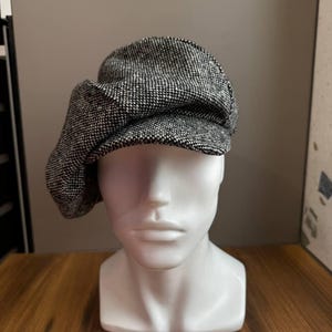 May include: A black and white tweed newsboy cap. The cap is displayed on a white mannequin head. The hat has a rounded crown and a small, stiff brim. The fabric has a textured appearance.