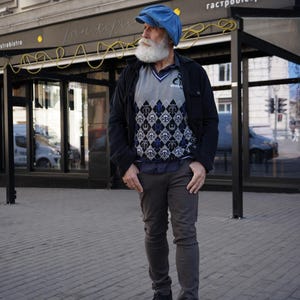 May include: A man with a long white beard is wearing a blue newsboy cap, a black jacket, a gray sweater with a geometric pattern, gray pants, and black shoes. He is standing in front of a building with a sign that says "гастробистро".
