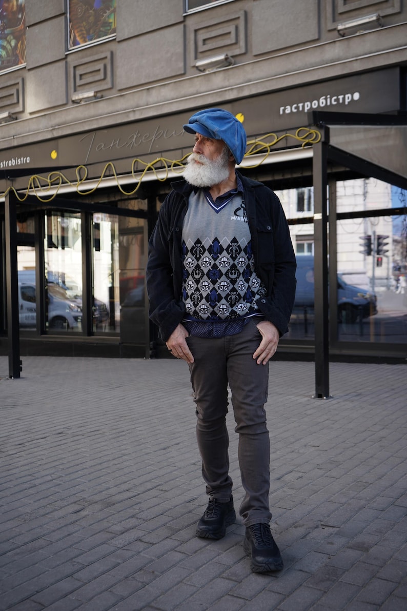 May include: A man with a long white beard is wearing a blue newsboy cap, a black jacket, a gray sweater with a geometric pattern, gray pants, and black shoes. He is standing in front of a building with a sign that says "гастробистро".