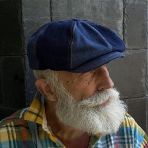 Puede incluir: Una gorra de estilo newsboy en mezclilla azul oscuro con una construcción de múltiples paneles y costuras naranjas visibles. La gorra presenta una corona redondeada y una visera corta y rígida en la parte frontal, hecha de algodón resistente.